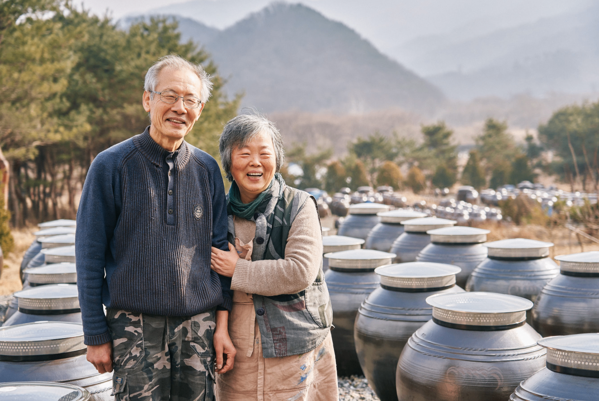Traditional Korean pottery artisan couple with ceramic vessels and mountain backdrop